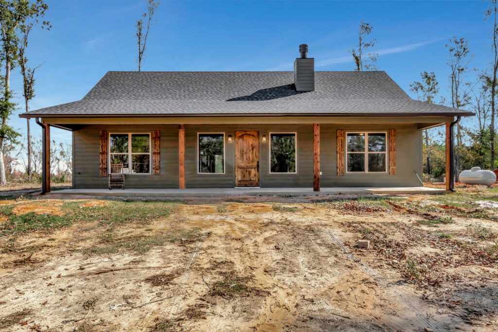 Single-story barndominium with a large covered front porch, wood shutters, and rustic wood columns, set in a wooded area with a clear blue sky.